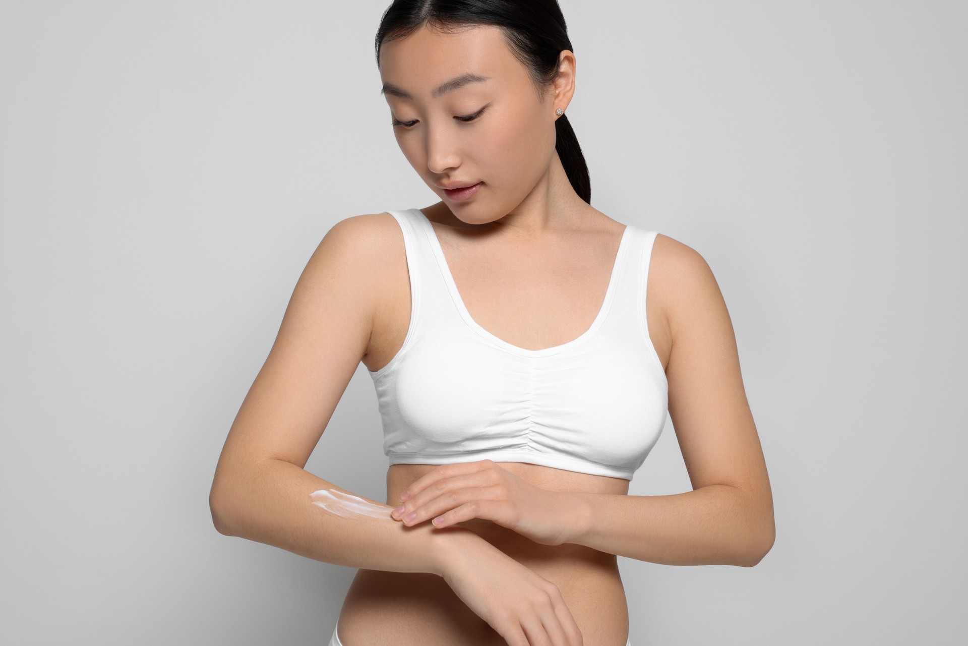 Beautiful young Asian woman applying body cream onto arm on light grey background