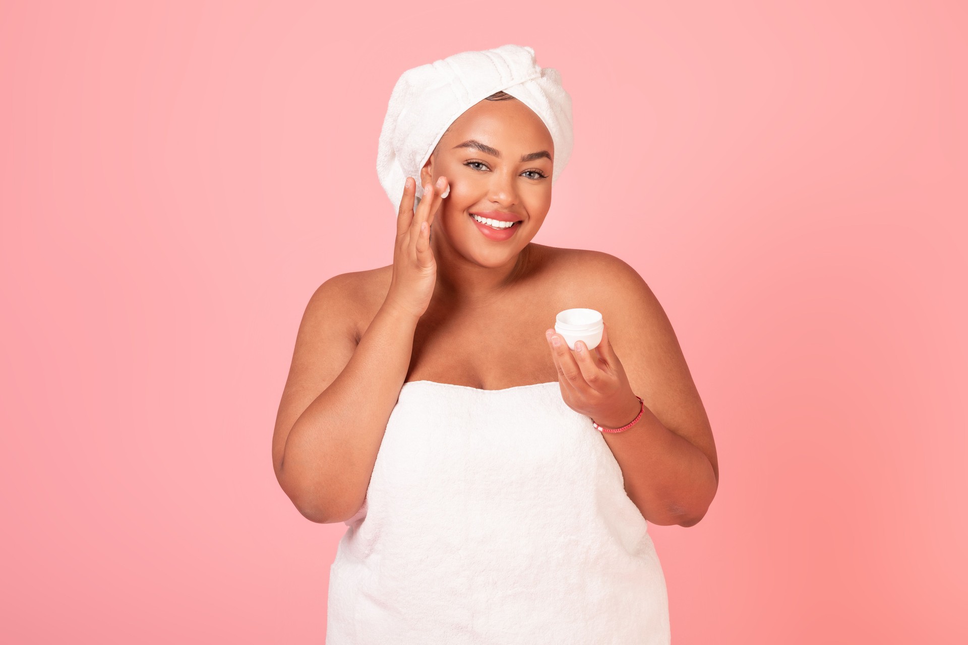 Skincare routine concept. Beautiful black chubby woman holding jar with moisturizing cream and applying on cheek