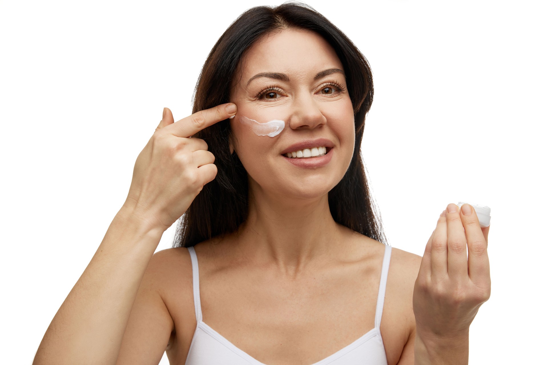 Portrait of attractive middle-aged woman applying facial cream against white studio background. Pleasure from self-care